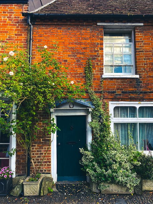 A close-up view of a traditional brick house with a dark green wooden front door, surrounded by lush, well-maintained garden plants. On the left, a tall flowering shrub with white blossoms and green f