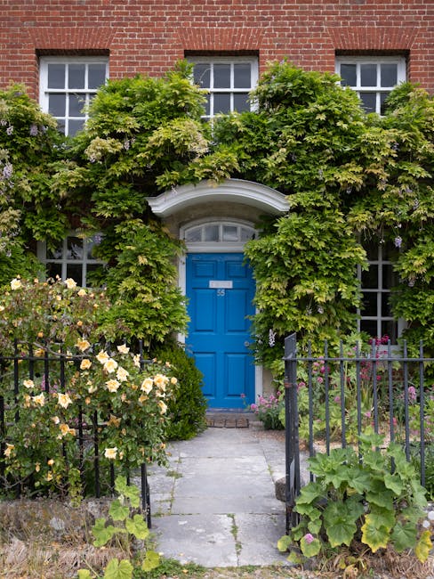 A charming front garden featuring a vibrant blue door set within a red-brick building. The entrance is framed by lush green foliage and climbing plants, including ivy and flowering vines, which partia